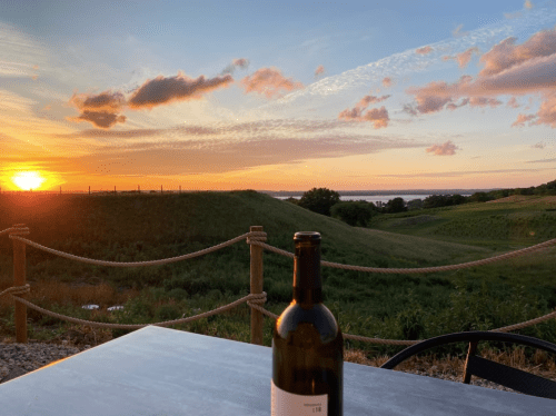 A bottle of wine sits on a table with a scenic sunset over rolling hills and a river in the background.