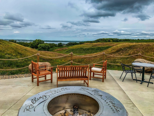 A scenic view of rolling hills and a lake, featuring outdoor seating and a fire pit under a cloudy sky.