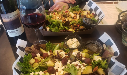 Two salads with mixed greens, fruits, nuts, and cheese, accompanied by a glass of red wine and a bottle on the table.