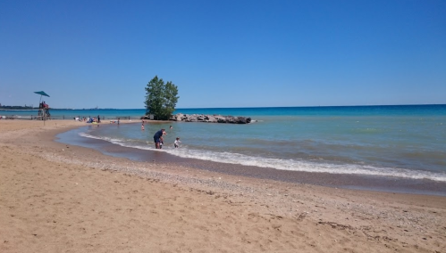 A sunny beach scene with people playing in the water and a clear blue sky over a calm lake.