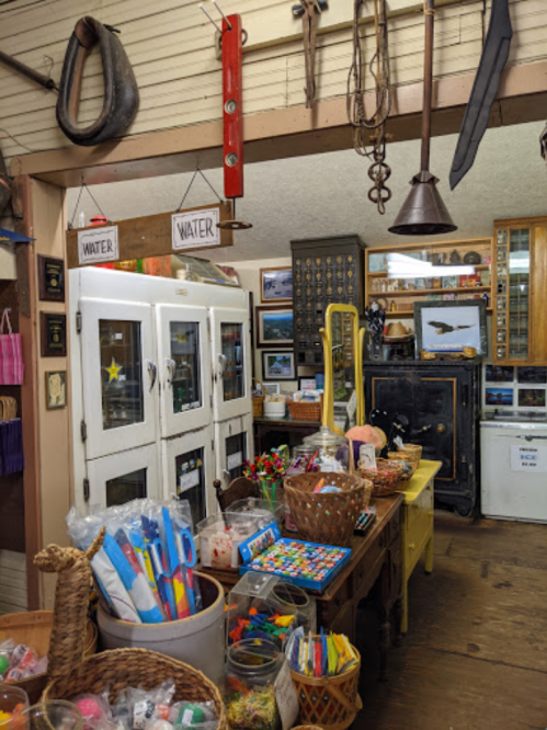 A cozy shop interior filled with colorful craft supplies, vintage decor, and display cases. Signs labeled "WATER" hang above.