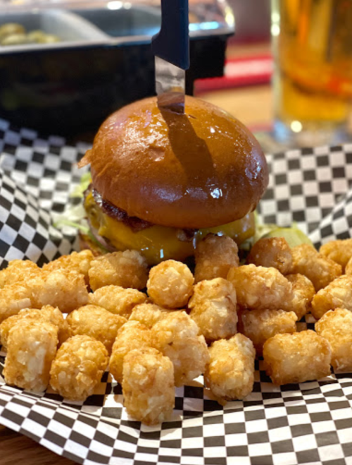 A juicy cheeseburger with a knife on top, surrounded by crispy tater tots on a checkered paper plate.
