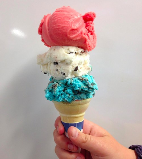 A hand holding a triple scoop ice cream cone with pink, white, and blue flavors against a white background.