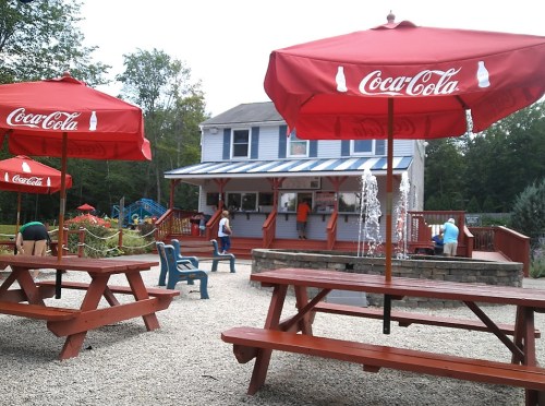 A cozy outdoor area with red Coca-Cola umbrellas, picnic tables, and a building in the background surrounded by trees.