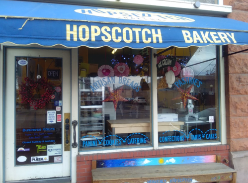 A bakery storefront with a blue awning, displaying "Hopscotch Bakery" and signs for artisan breads and pastries.