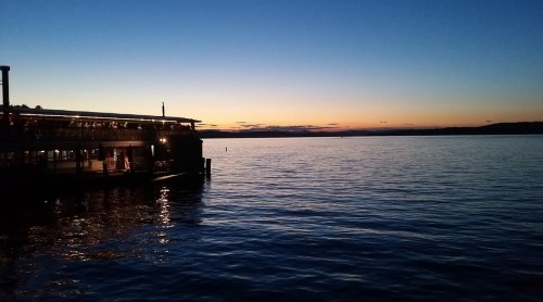A serene sunset over calm water, with a silhouette of a docked building on the left and distant hills on the horizon.