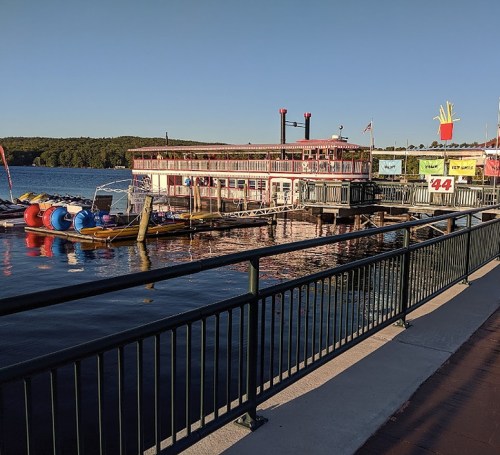 A waterfront scene featuring a restaurant boat, colorful watercraft, and a clear blue sky reflecting on the water.