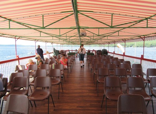 Interior of a ferry with empty brown chairs, a striped canopy, and a view of water in the background.