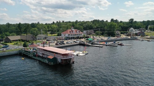 Aerial view of a waterfront area with a boat dock, restaurants, and green trees under a partly cloudy sky.