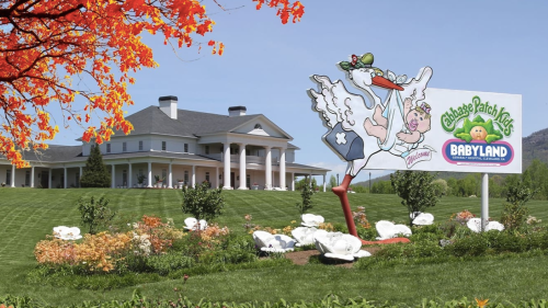 A colorful sign for Cabbage Patch Kids Babyland in front of a large white mansion and vibrant landscaping.