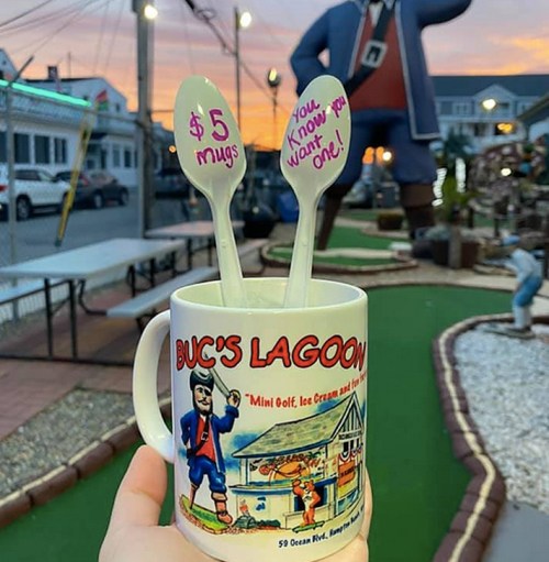 A hand holds a colorful mug with "Buc's Lagoon" design, featuring spoons and a mini golf course in the background at sunset.