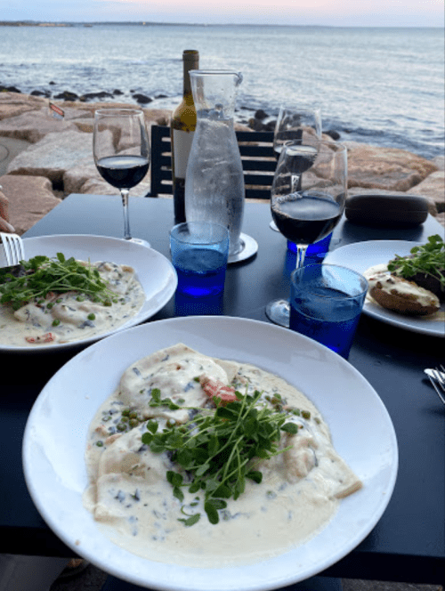 A seaside dining table with three plates of pasta in creamy sauce, wine glasses, and a pitcher of water.
