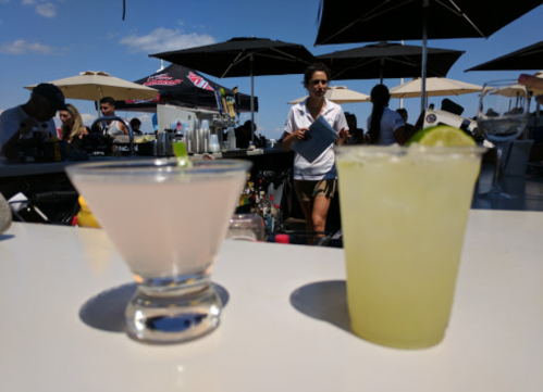 Two cocktails on a bar counter, with a bartender in the background and umbrellas providing shade.
