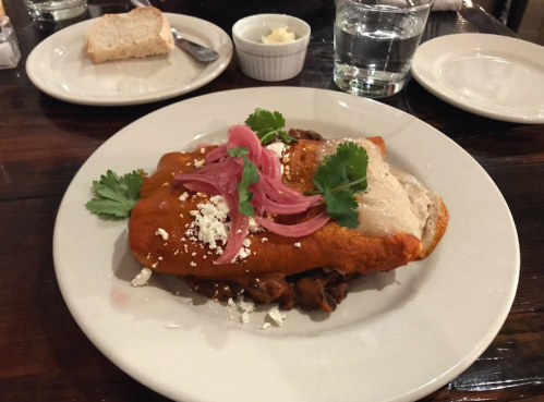 A plate of enchiladas topped with red sauce, cheese, pickled onions, and cilantro, served with a side of bread and water.