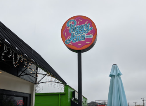 Sign for Peggy Ann Bakery featuring colorful lettering and a donut design, with a cloudy sky in the background.