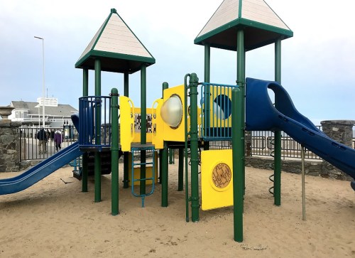 Colorful playground structure with slides and climbing features, set on sandy ground under a cloudy sky.