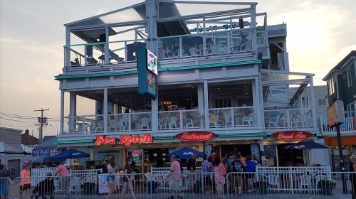 A multi-level beachside restaurant with outdoor seating, featuring a sign for "Sea Ketch" and a lively atmosphere.