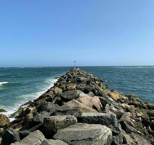 A rocky jetty extends into the ocean under a clear blue sky, with gentle waves lapping at the stones.