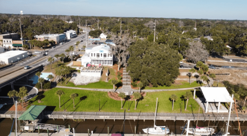 Aerial view of a park with green grass, palm trees, and a waterfront, surrounded by buildings and roads in the background.