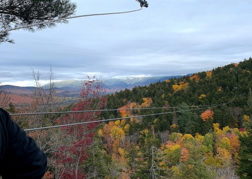 A scenic view of colorful autumn foliage on mountains under a cloudy sky, with zip line cables in the foreground.