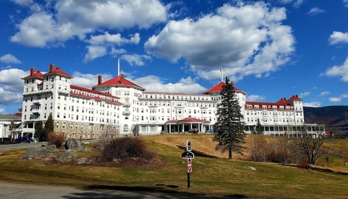 A large, historic hotel with a white facade and red roofs, set against a blue sky with fluffy clouds and green grass.