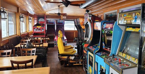 A cozy arcade room with various vintage arcade machines and empty tables and chairs.