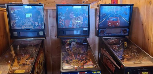 Three vintage pinball machines lined up against a wooden wall, featuring colorful graphics and illuminated screens.
