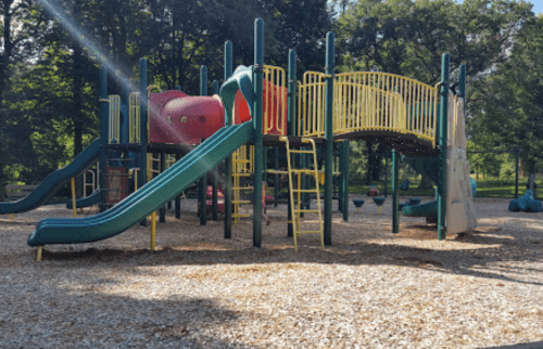 Colorful playground equipment with slides and climbing structures, surrounded by trees and wood chip ground cover.