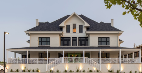 A large, elegant two-story house with a wraparound porch and modern architectural details, set against a twilight sky.