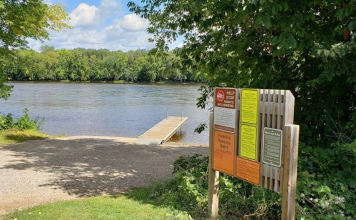 A riverbank scene with a wooden dock and informational signs about invasive species and conservation efforts.
