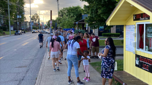 A busy street scene with a diverse group of people walking past a yellow ice stand on a sunny day.