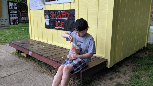 A child sits on a wooden platform outside a yellow shack, enjoying a bowl of shaved ice on a sunny day.