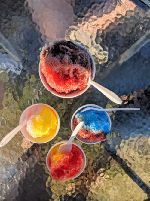 Four colorful bowls of shaved ice in various flavors sit on a glass table, with spoons beside them.