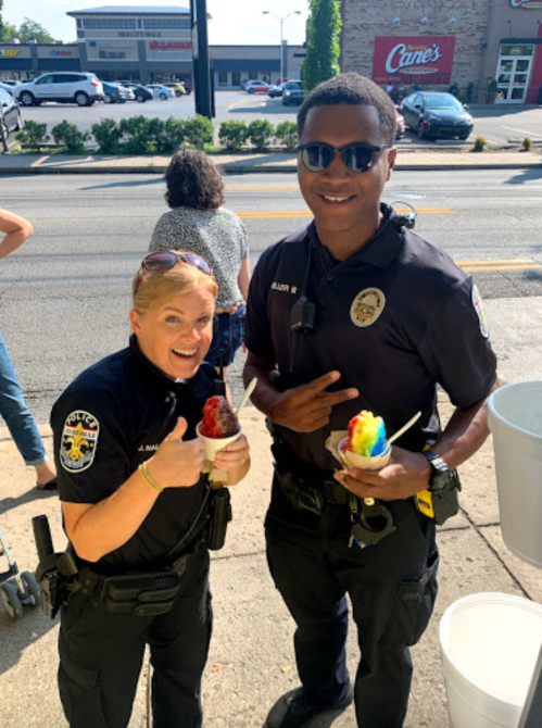 Two police officers smiling and holding cups of colorful shaved ice, standing outdoors on a sunny day.