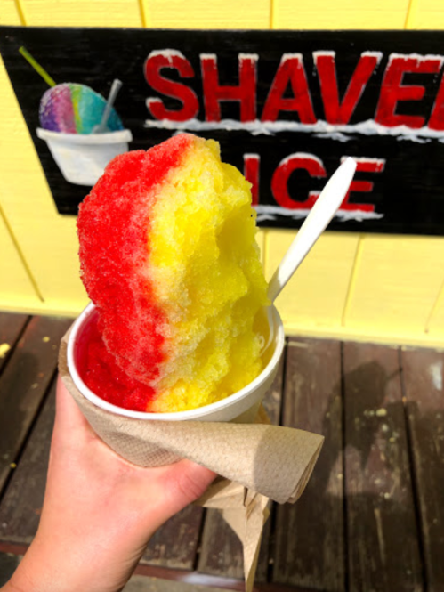 A hand holding a cup of colorful shaved ice with red and yellow layers, set against a bright yellow wall.