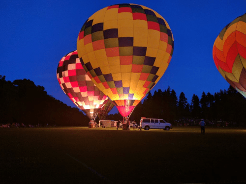 Colorful hot air balloons illuminated at dusk, with a crowd in the background and a vehicle nearby.