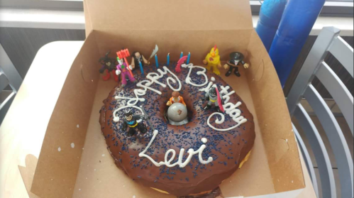 A large chocolate donut in a box, decorated with "Happy Birthday Levi" and colorful toy figures on top.