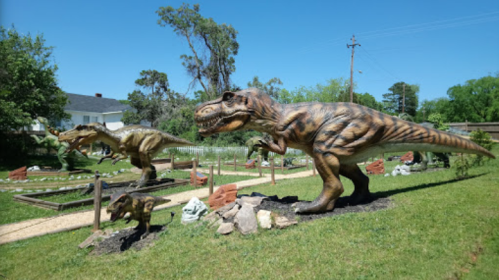 Three dinosaur sculptures, including two T. rex figures, in a grassy outdoor setting with trees and a building in the background.