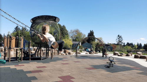A child swings at a playground on a sunny day, with another child swinging in the background and trees in the distance.