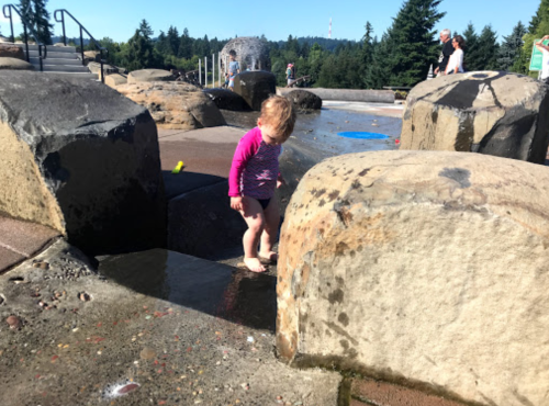A young child in a swimsuit stands barefoot between large rocks at a water play area, enjoying the splashing water.