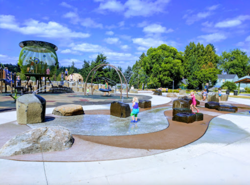 A vibrant playground with water features, rocks, and children playing under a blue sky with fluffy clouds.