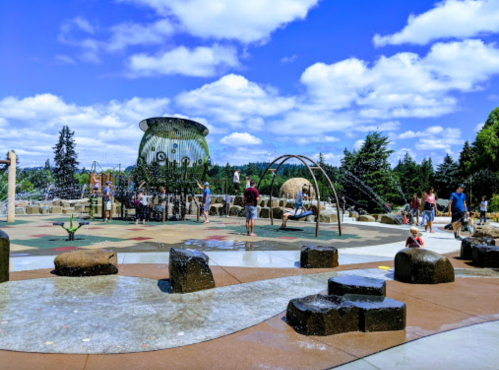 A vibrant playground with water features, children playing, and a blue sky with fluffy clouds in the background.