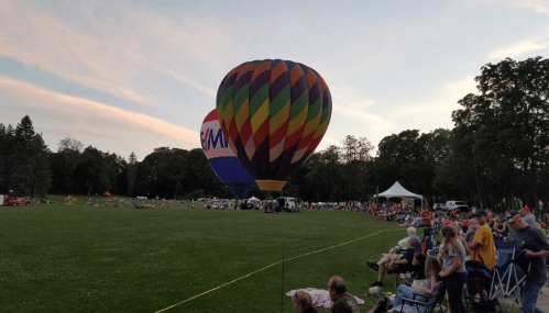 Two colorful hot air balloons inflate at dusk, surrounded by a crowd on a grassy field with trees in the background.