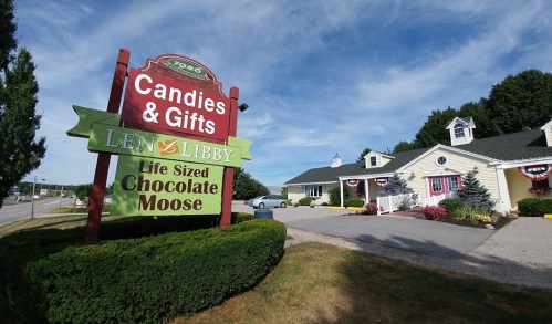 Sign for "Len & Libby Candies & Gifts" featuring a life-sized chocolate moose, with a charming building in the background.