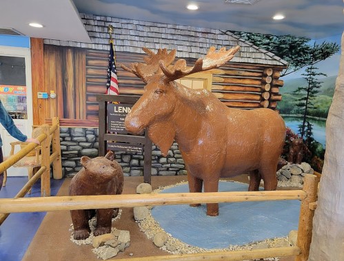A life-sized moose sculpture stands next to a bear sculpture in a rustic indoor setting with a wooden fence.