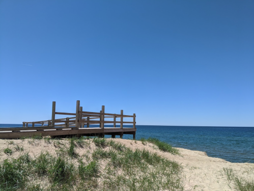 A wooden dock extends over a sandy beach, with clear blue water and a cloudless sky in the background.