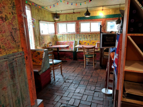 Cozy restaurant interior with colorful decor, wooden furniture, and brick flooring, featuring string lights and artwork on walls.