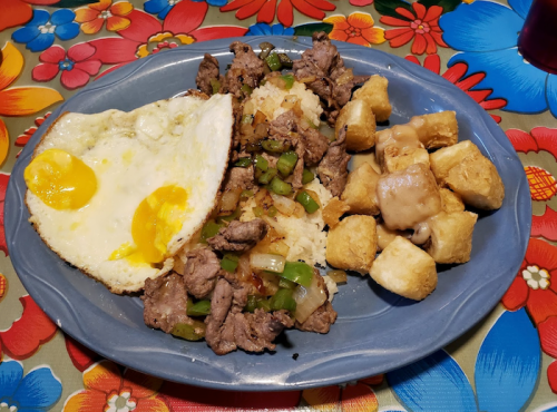 A blue plate with steak, scrambled eggs, rice with peppers, and crispy potato bites drizzled with sauce on a floral tablecloth.