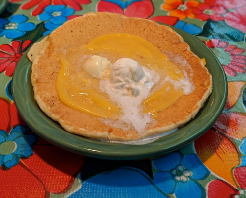 A large pancake topped with melted butter and a scoop of ice cream, served on a colorful floral tablecloth.