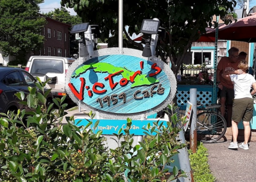 Sign for Victor's 1959 Café, featuring a colorful design and surrounded by greenery and outdoor seating.